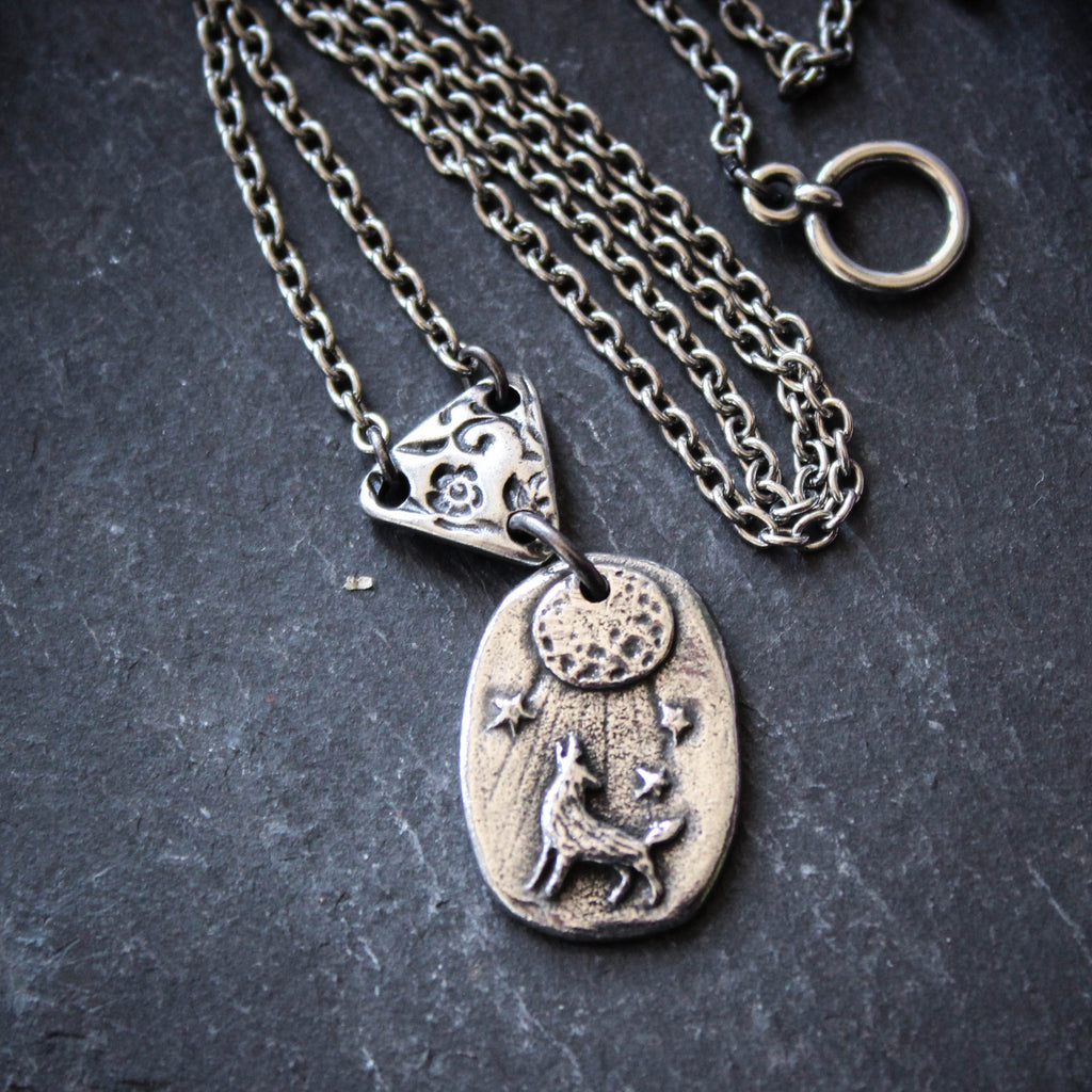 A silver and bronze necklace featuring a small wolf medallion with a glowing full moon and stars, connected to a chain made of recycled silver.
