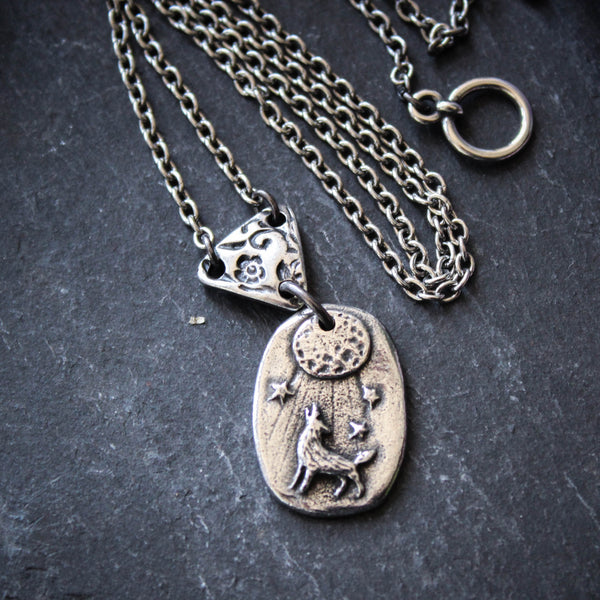 A silver and bronze necklace featuring a small wolf medallion with a glowing full moon and stars, connected to a chain made of recycled silver.